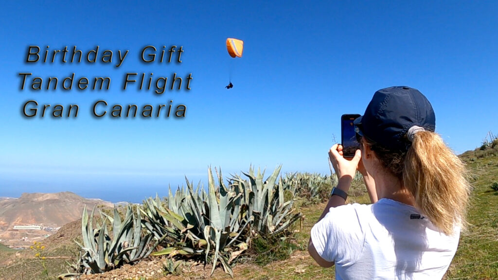 A tandem paragliding flight over Gran Canaria with a birthday girl soaring through the sky while her friend records the moment from the ground. The paraglider glides above stunning coastal views, capturing an unforgettable birthday adventure